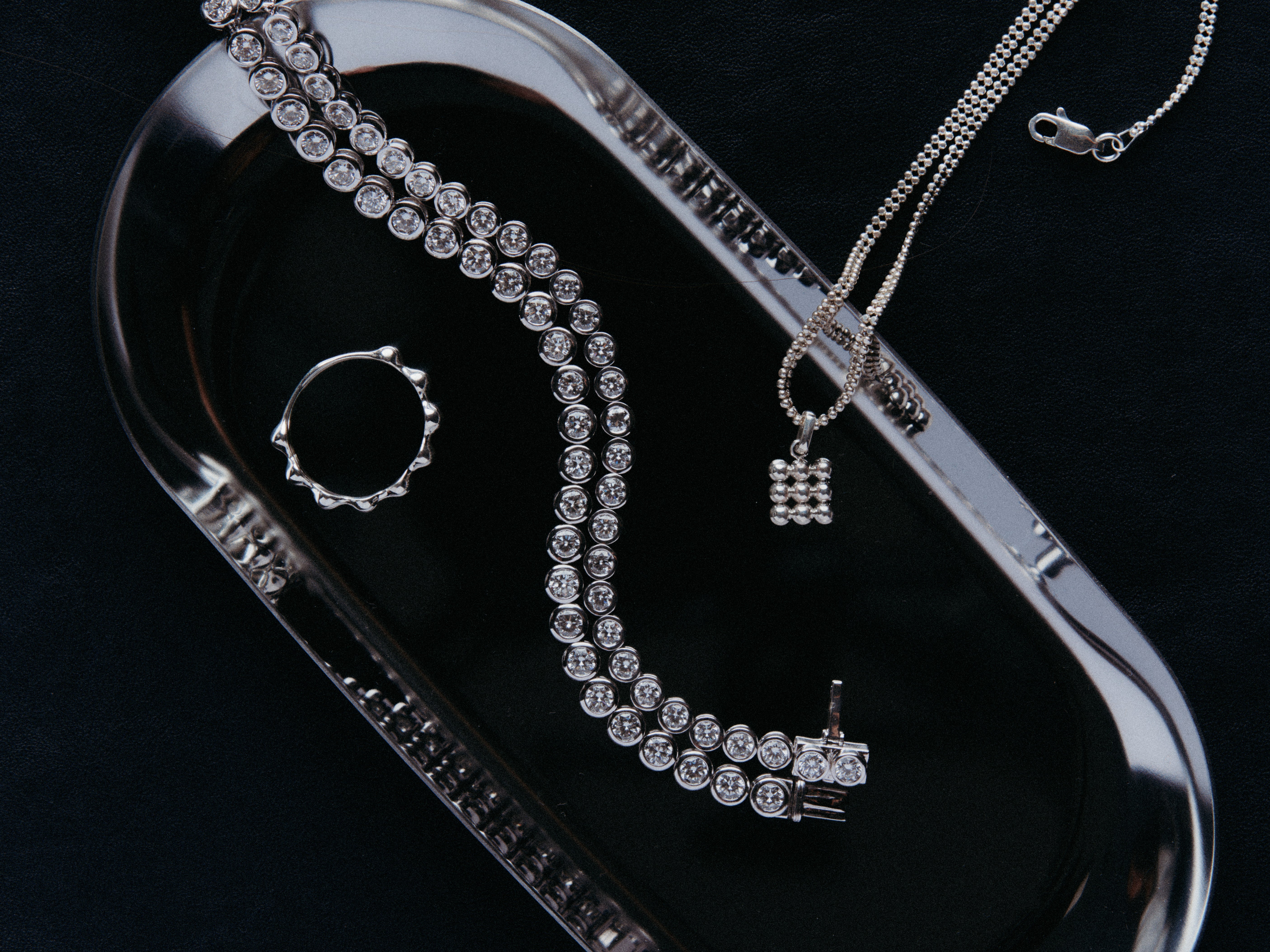 Axis Ring, Roller Bracelets, Grid Pendant and Double Row Chain on a reflective metal plate sitting on a dark background.